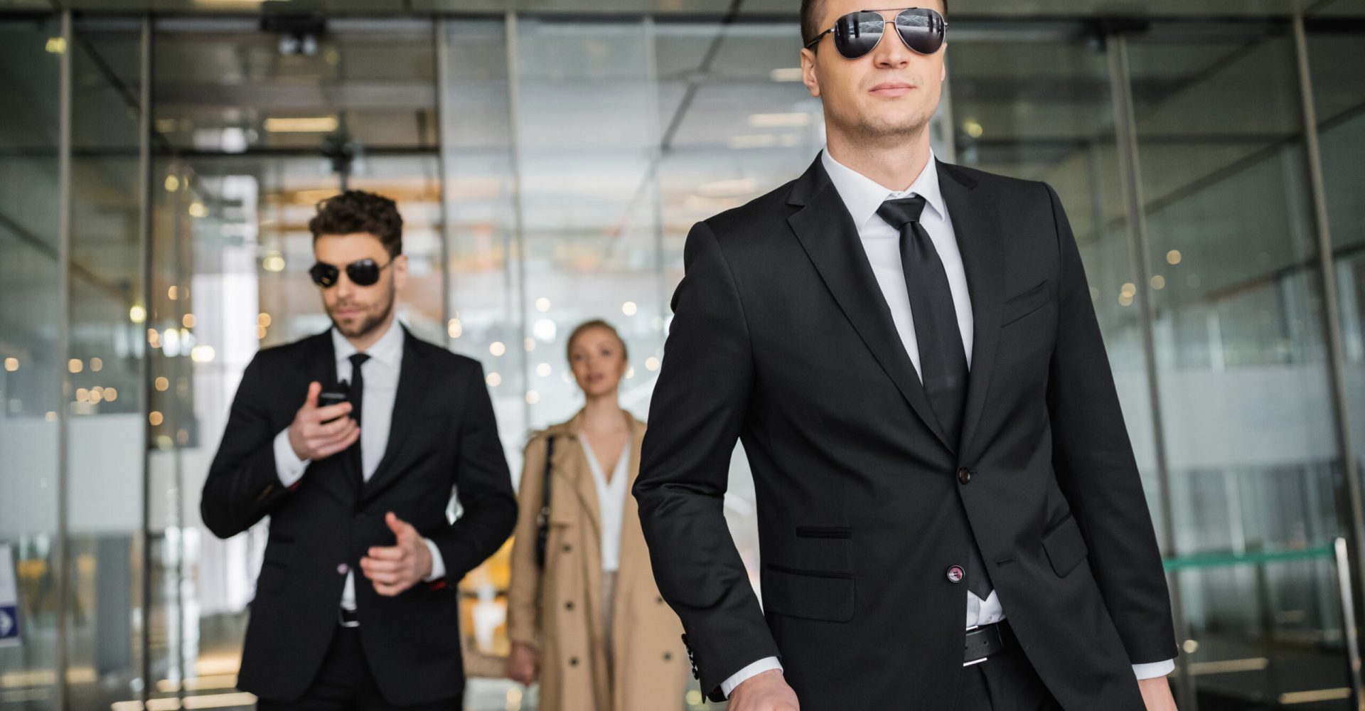 private security service, personal safety concept, handsome bodyguards in formal wear and sunglasses protecting safety of blonde client on blurred background, men and woman walking in hotel